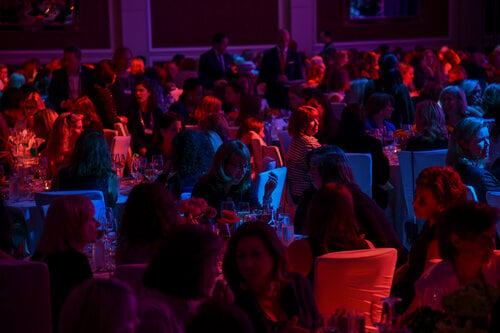 Large crowd at an evening event, seated at round tables, chatting under dim, colorful lighting.